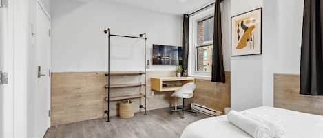 A modern hotel room with a wooden floor, a bed with white linens, a desk with a chair, a hanging towel rack, and a cityscape view through the window.