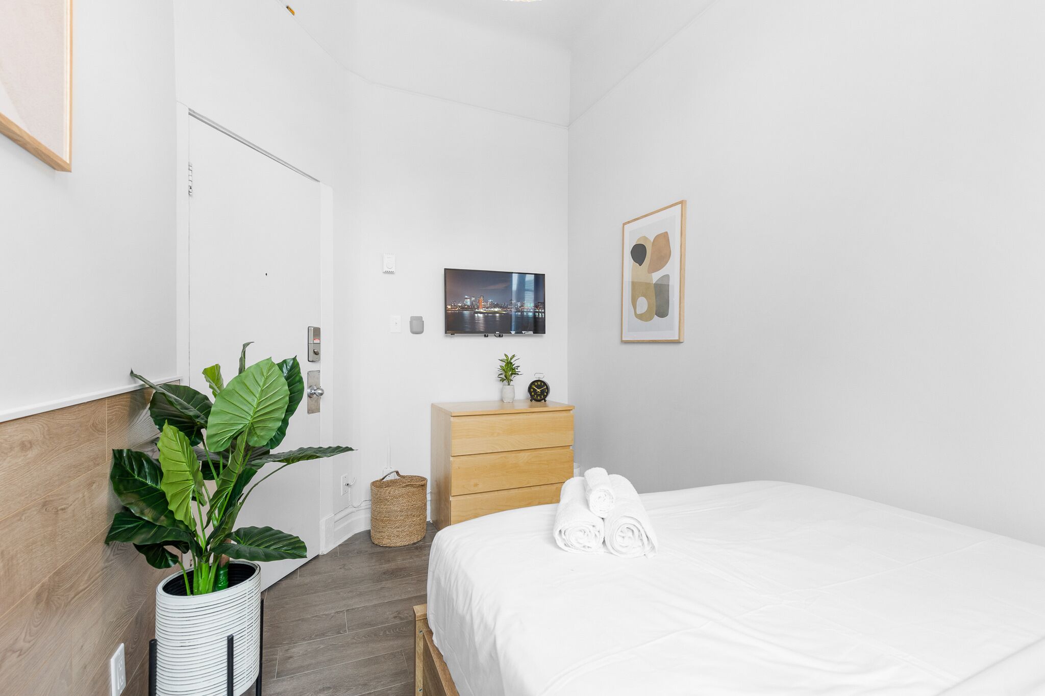 Habitación de hotel moderna con cabecera de madera, una planta grande en maceta, cómoda de madera, cama con ropa de cama blanca y un cuadro en la pared.