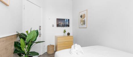 Habitación de hotel moderna con cabecera de madera, una planta grande en maceta, cómoda de madera, cama con ropa de cama blanca y un cuadro en la pared.