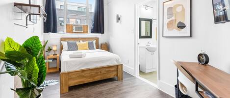 A modern bedroom with a wooden bed, a large potted plant, a hanging light fixture, and a view of the city through the window.