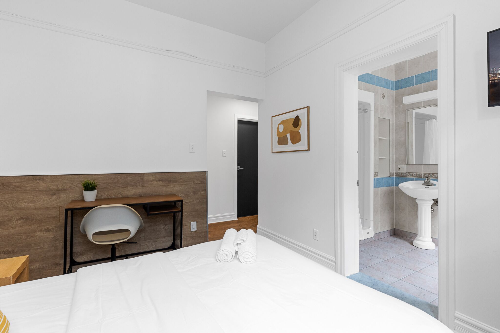 A hotel room with a bed, a desk with a chair, a small plant, and a bathroom with a sink and mirror.