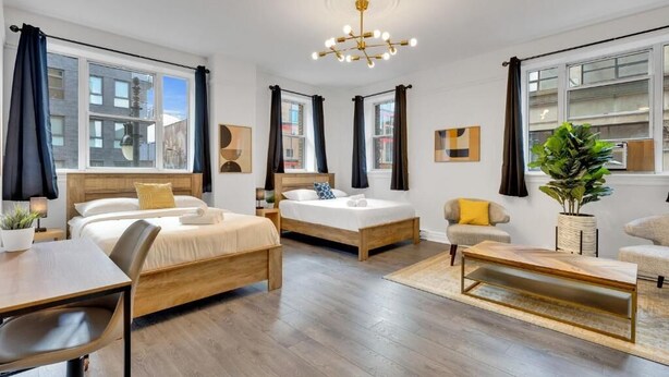 A modern hotel room with two beds, a dining table, a sofa, and a large window with curtains. - Le Sand & Stone Hôtel (Montreal)