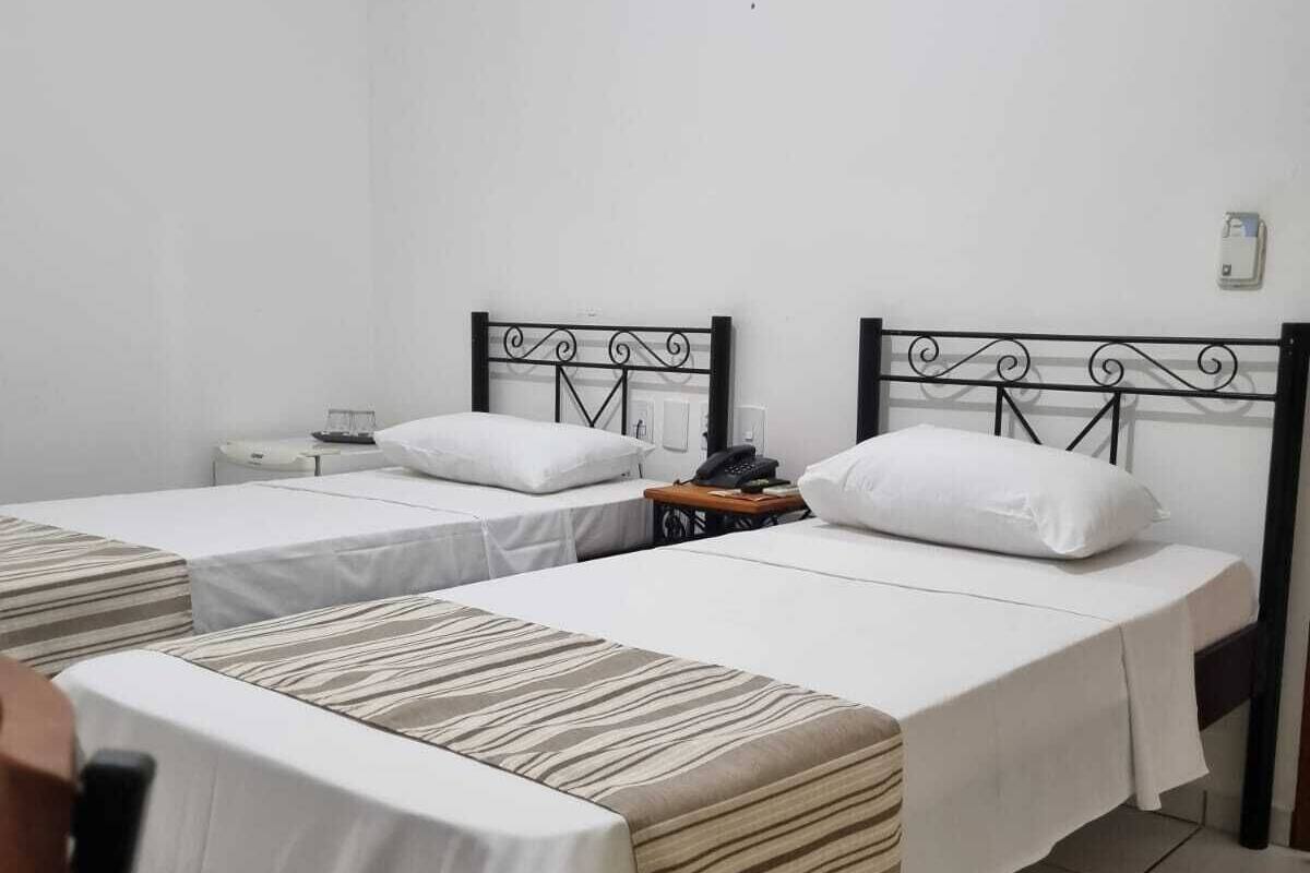 Una habitación de hotel con dos camas individuales, una mesita de noche pequeña con lámpara y una pared blanca lisa.