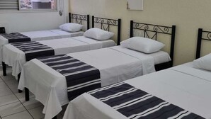 Une chambre d’hôtel avec quatre lits simples, chacun recouvert d’une couvre-lit à rayures et agrémenté d’oreillers blancs. La chambre dispose d’une fenêtre donnant sur la ville, d’un climatiseur fixé au mur et d’un petit objet décoratif placé au-dessus des lits.