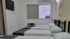 Une chambre d’hôtel avec trois lits, un téléviseur fixé au mur, un climatiseur placé au-dessus de la fenêtre et une vue sur le paysage urbain.