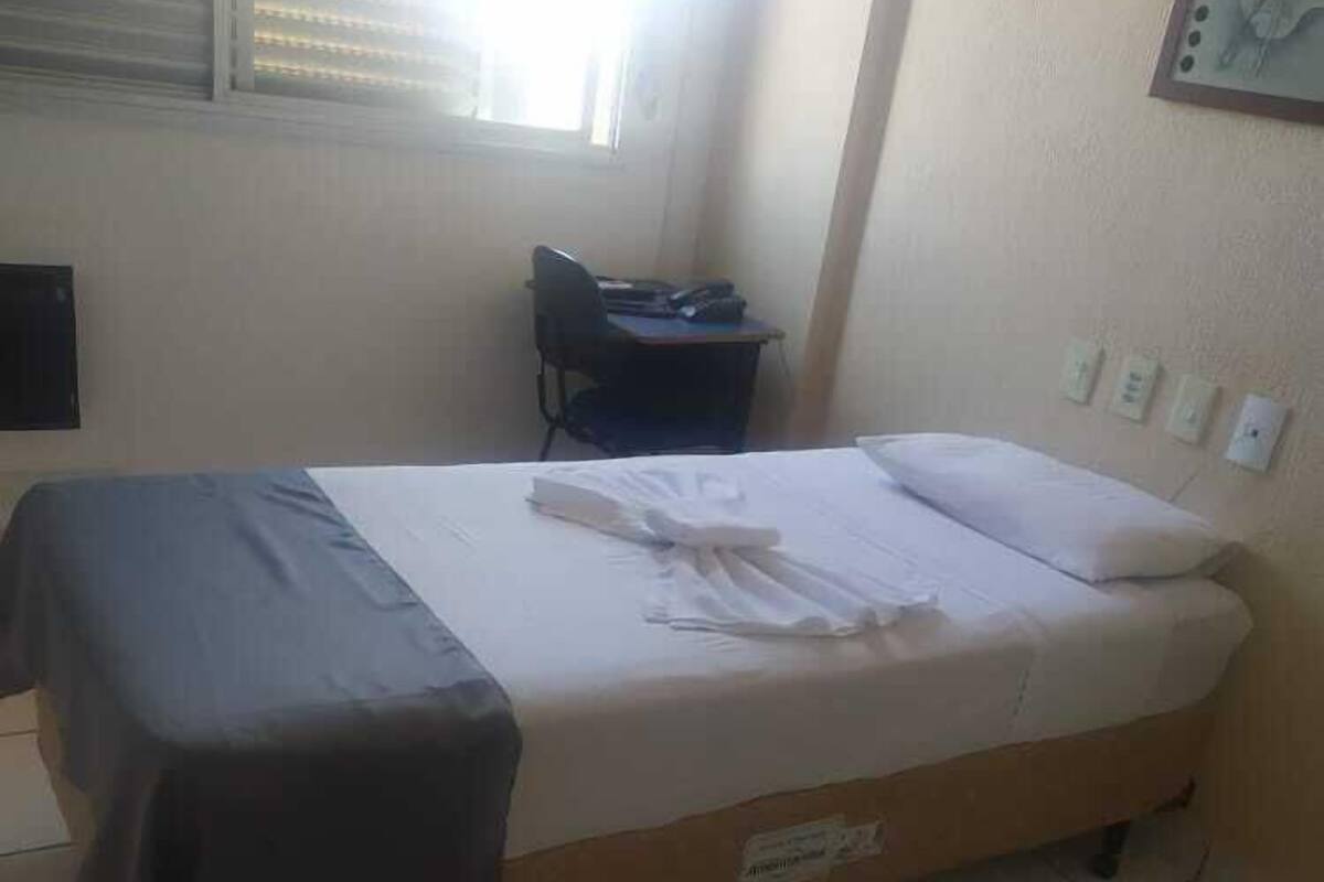 Una cama individual con colchón blanco y cabecera marrón, un escritorio con silla, y una ventana con persianas.