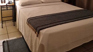 Un lit bien fait, recouvert d’une courtepointe blanche et agrémenté d’oreillers, une table de chevet avec une lampe, et une petite table avec une chaise.
