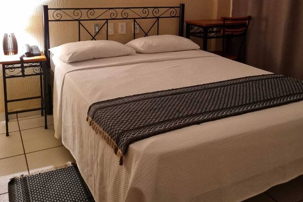 Una cama bien hecha con colcha y almohadas blancas, una mesita de noche con lámpara, y una pequeña mesa con una silla.