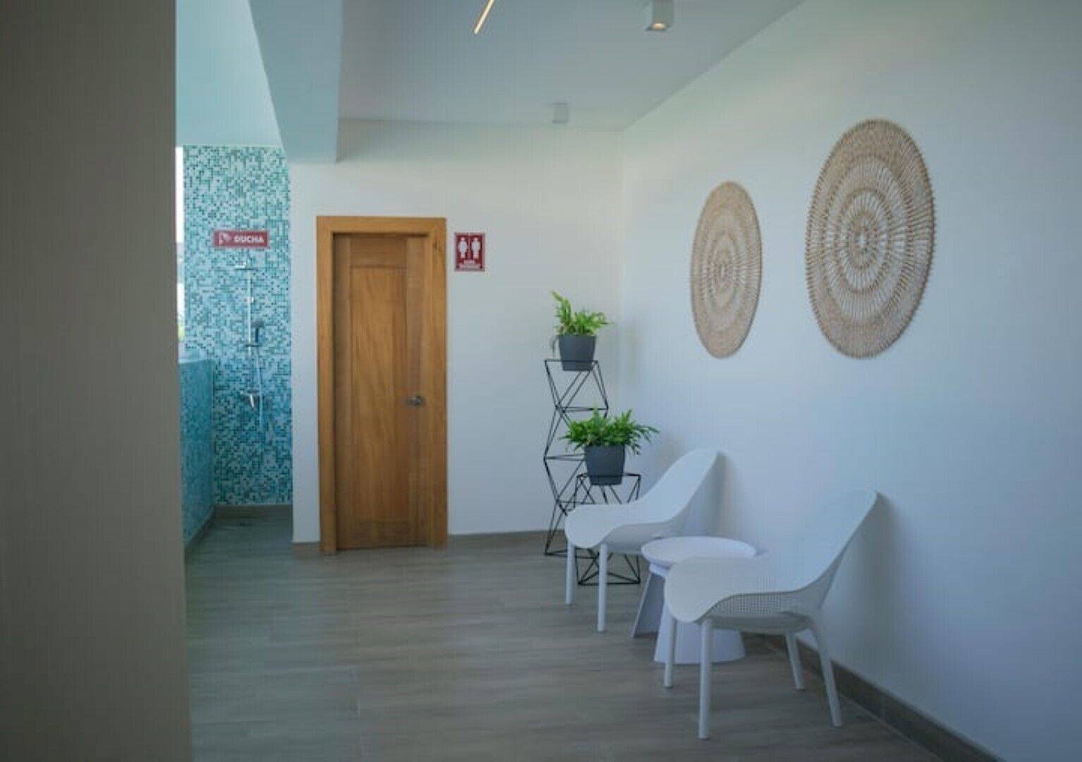 Un couloir avec une porte en bois, un comptoir d’accueil, deux chaises blanches et un présentoir à plantes avec des plantes en pot.