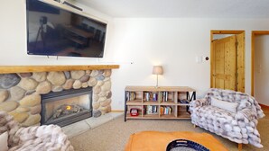 A cozy living room with a stone fireplace, a television mounted above it, a bookshelf, and a plush armchair. - Welcome to Cozy at the Crystal (Sun Peaks)