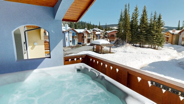 A hot tub with a wooden deck and a view of snow-covered buildings and trees. - Welcome to Cozy at the Crystal (Sun Peaks)