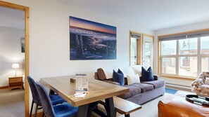Una habitación con una mesa de comedor de madera, sillas tapizadas en azul, un sofá gris con cojines, un ventanal con vistas a edificios y un cuadro en la pared.
