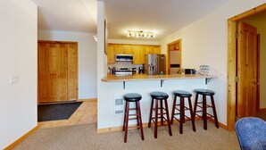 A compact kitchen with wooden cabinets, a central island with stools, and a refrigerator. - Welcome to Cozy at the Crystal (Sun Peaks)
