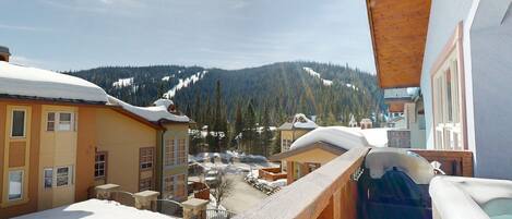 A snow-covered balcony with a hot tub, overlooking a ski resort with buildings and mountains.