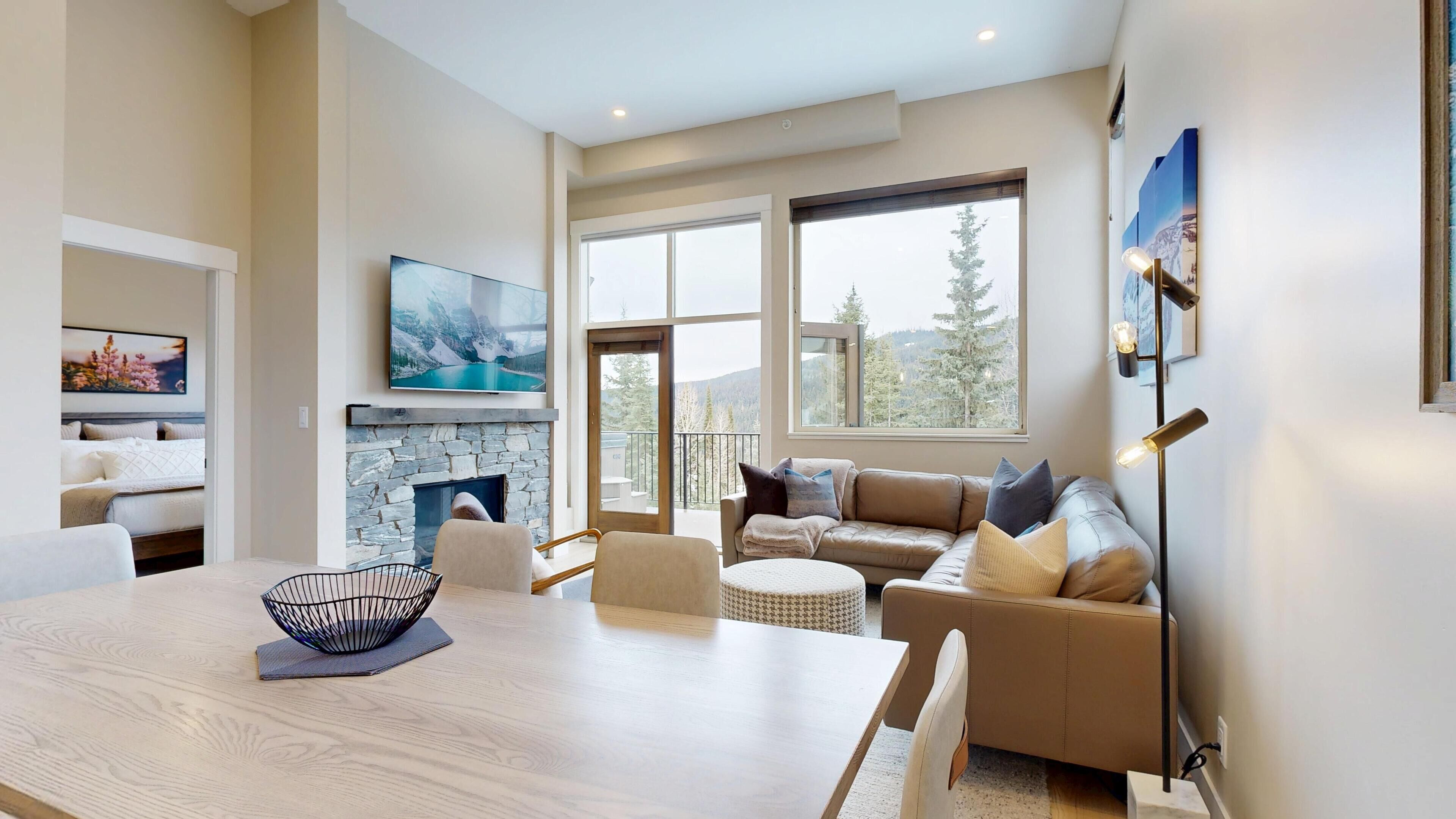 A modern living room with a stone fireplace, a large window with a view of mountains, a dining area with a wooden table, and a sofa with cushions.