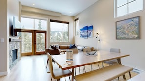 A modern living room with a fireplace, a dining table with chairs, a sofa, and a large window offering a view of trees and mountains.
