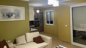 Una sala de estar con sofá, una araña de techo, una cocina al fondo y un ventanal con persianas.
