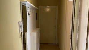 Un pasillo con una puerta blanca al final, una pared blanca con un teléfono y una lámpara colgante.