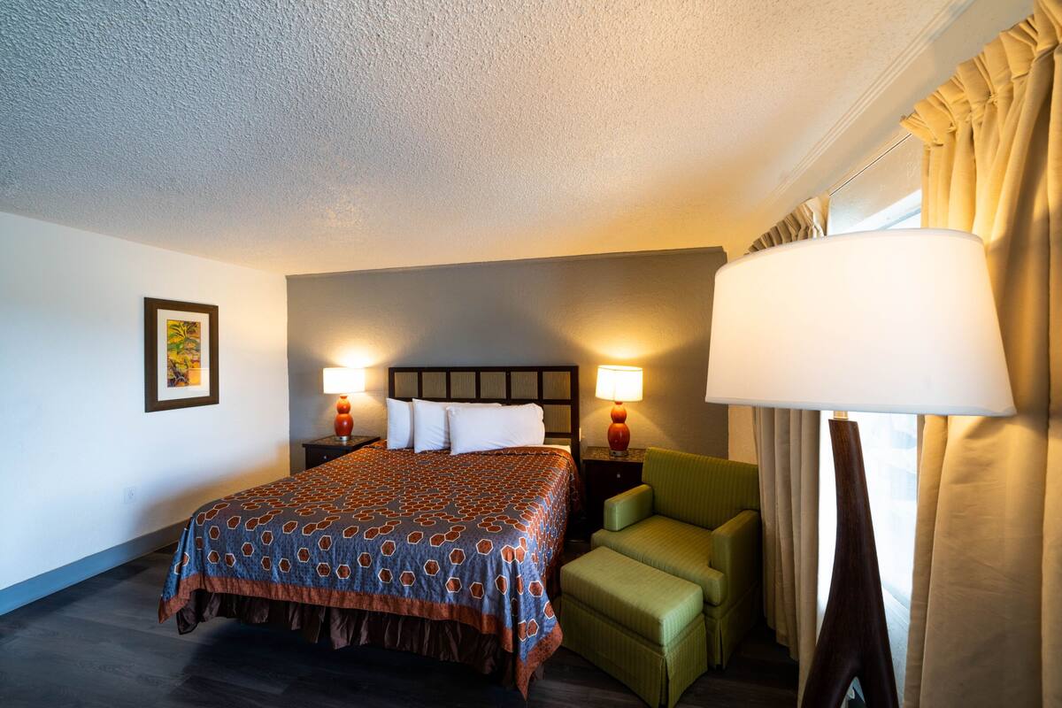 A hotel room with a bed, two bedside lamps, a green armchair, a large floor lamp, and a window with curtains.