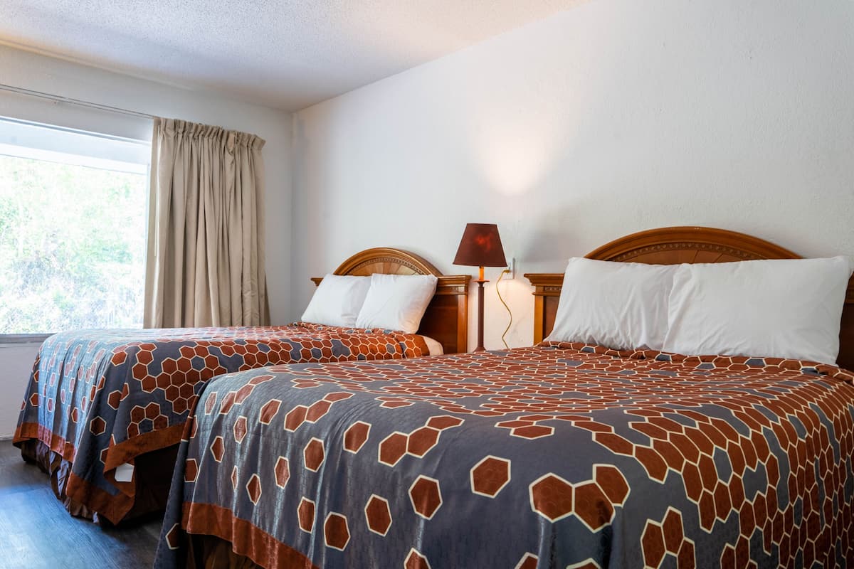 A hotel room with two beds, each with a patterned bedspread, a wooden headboard, a bedside table with a lamp, and a window with curtains.