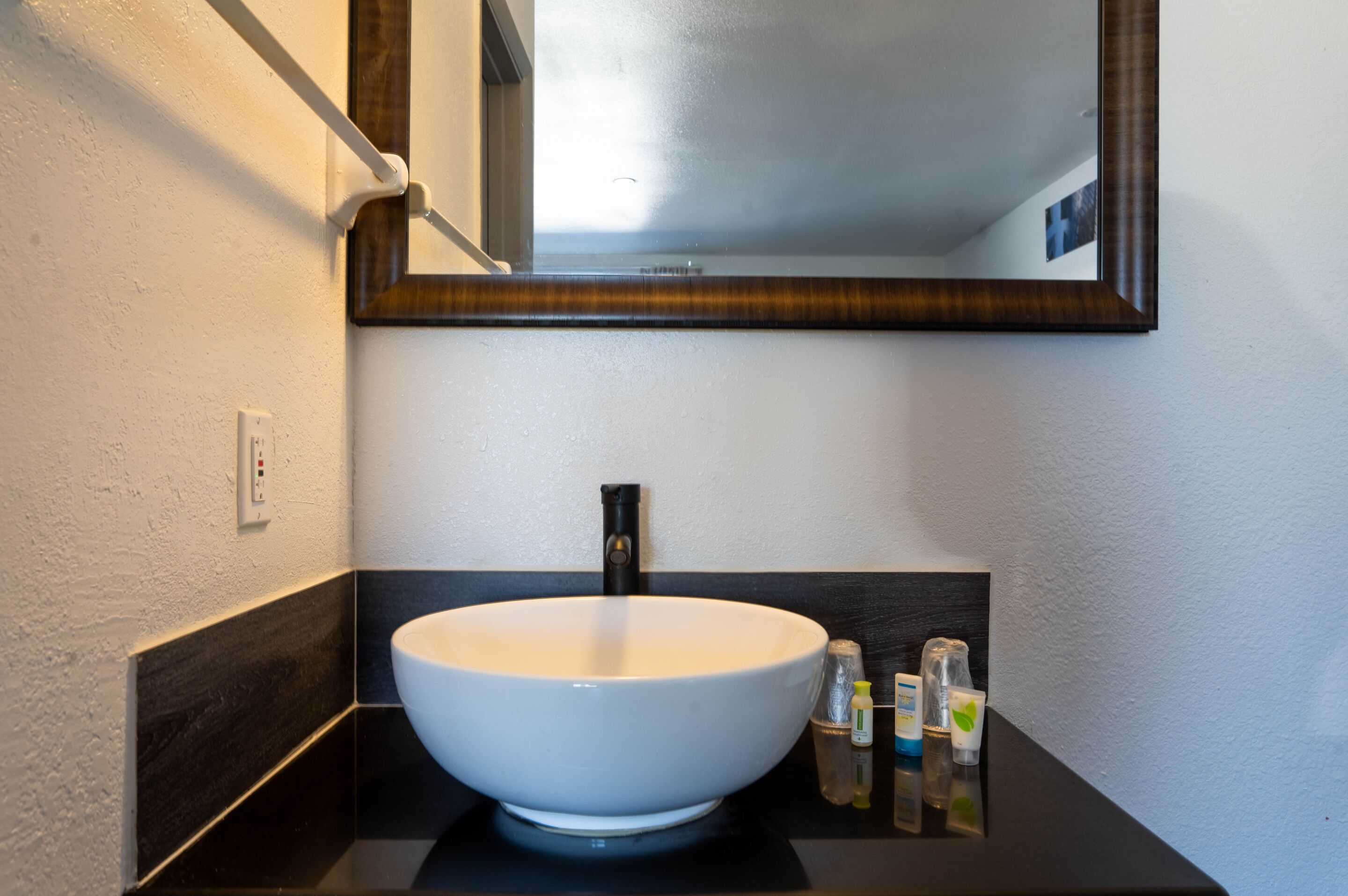 Un bagno moderno con un ampio lavabo bianco a incasso, un rubinetto nero e uno specchio con cornice in legno. Sul mobile del lavabo sono presenti articoli da toilette.