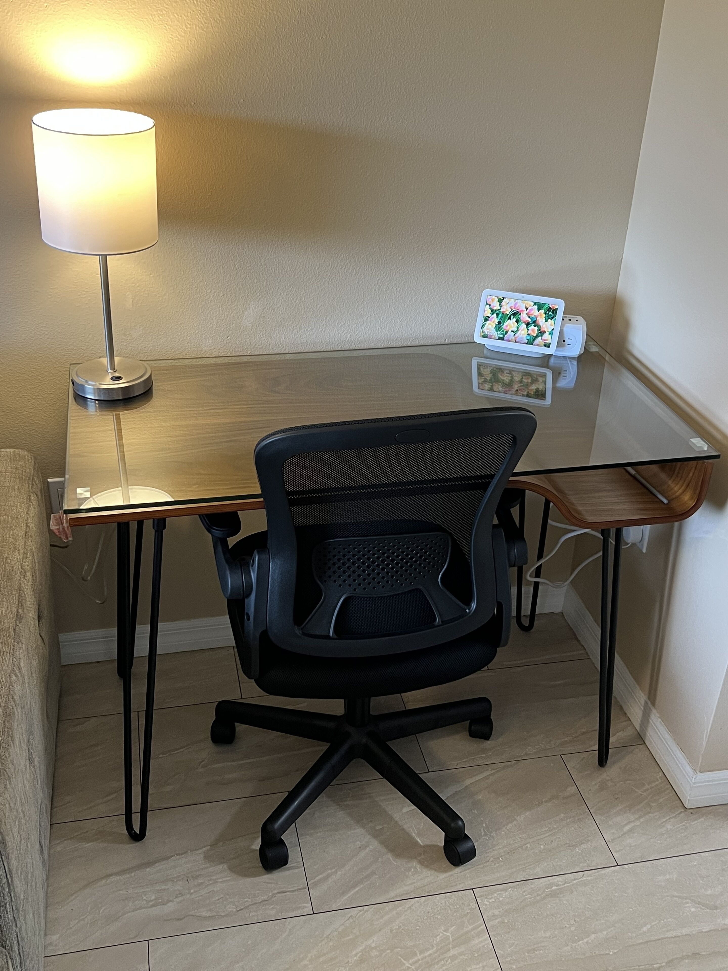 A small, modern office space with a glass table, a black office chair, and a lamp.