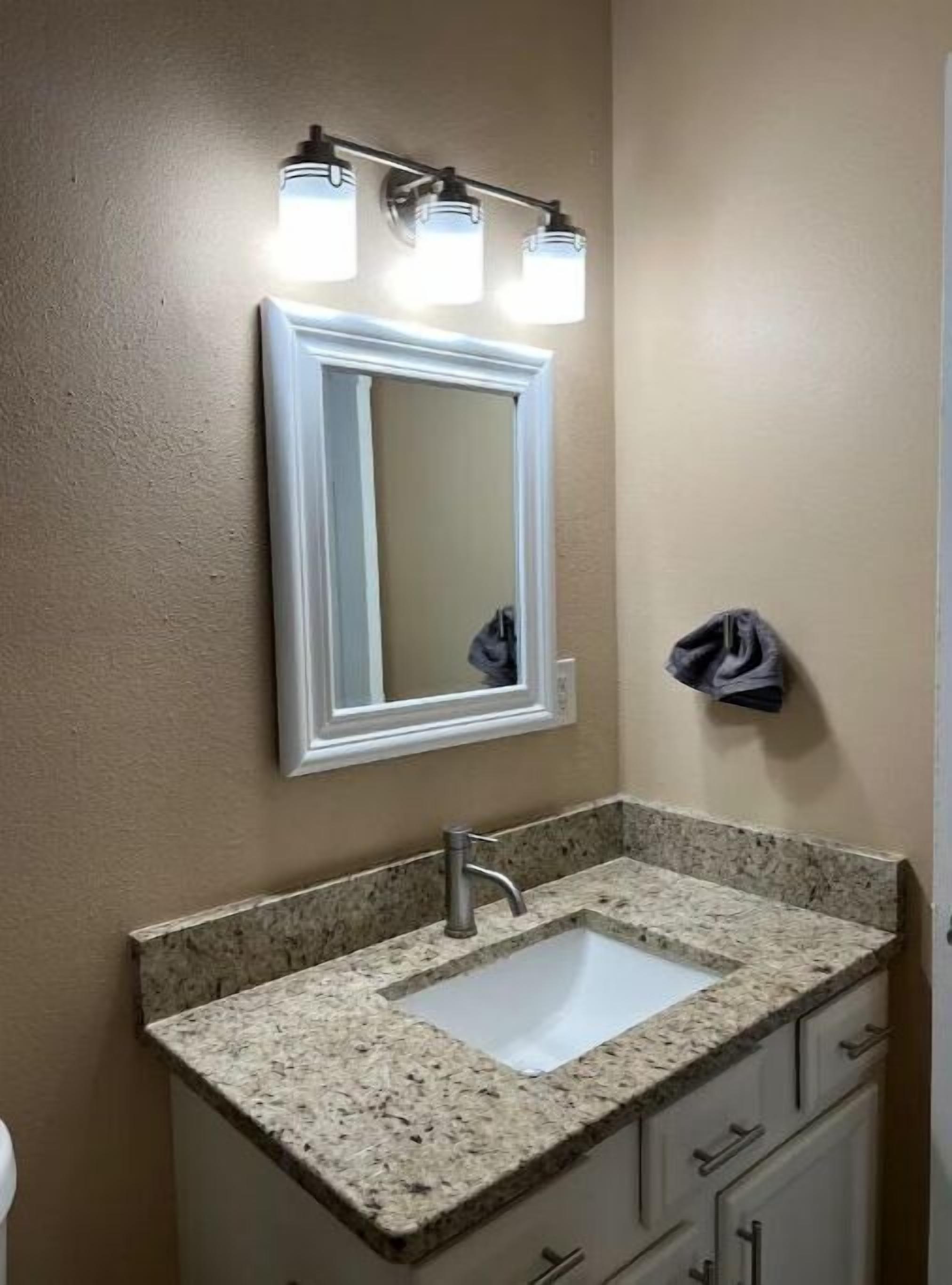 A bathroom with a granite countertop sink, a white vanity, and a mirror with three lights above it.