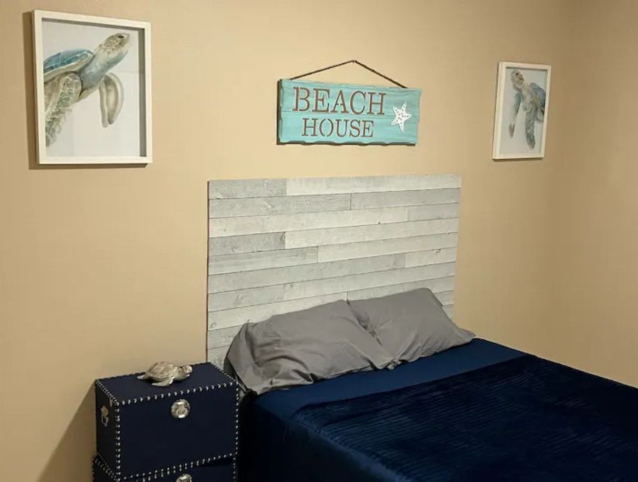A bedroom with a wooden headboard, a bed with grey bedding, a small bedside table with a decorative item, and two framed pictures of sea turtles on the walls.