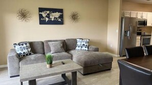 A modern living room with a sectional sofa, a coffee table, decorative wall art, and a kitchen area in the background. - Peace and Quiet near Daytona (Holly Hill)
