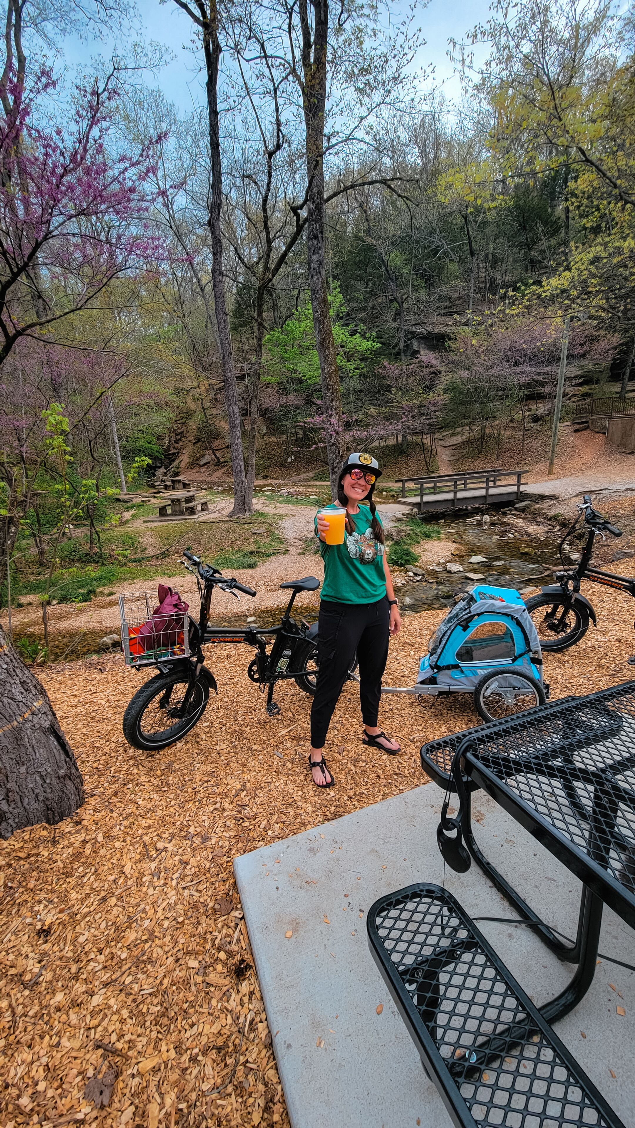 A person standing next to a bicycle and a picnic table in a park with trees and a picnic bench.