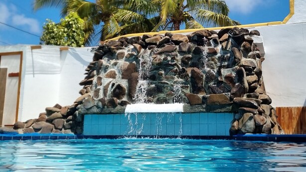 A swimming pool with a waterfall feature, surrounded by palm trees and a clear blue sky. - Fernandina Hotel & Spa Galápagos (Puerto Ayora)