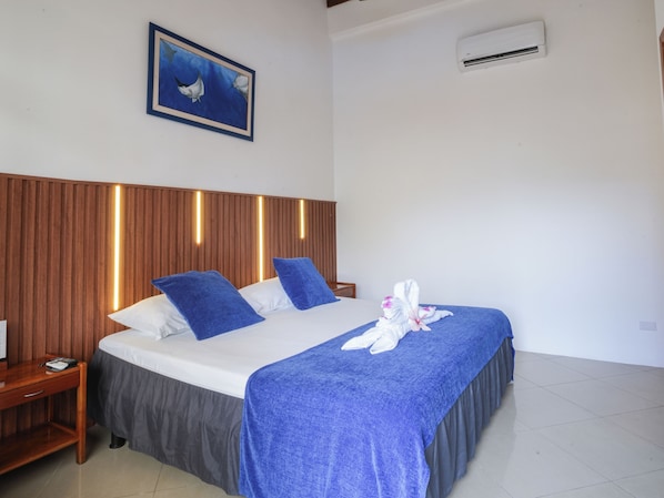 A hotel room with a wooden headboard, a bed with blue and white bedding, a blue throw blanket, and a towel with pink flowers. - Fernandina Hotel & Spa Galápagos (Puerto Ayora)