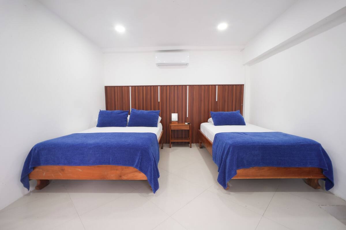 Ein Zimmer mit zwei Einzelbetten, beide jeweils mit Holzrahmen und blauer Bettwäsche. Dazwischen steht ein kleiner Nachttisch aus Holz, und an der weißen Wand ist ein Holzpaneel-Design zu sehen.
