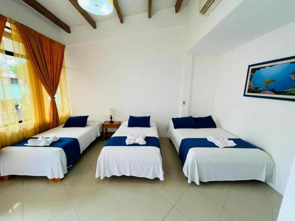Habitación de hotel con tres camas individuales, cada una con una colcha y almohadas blanco y azul. Hay una mesita con una lámpara entre las camas y un cuadro en la pared.