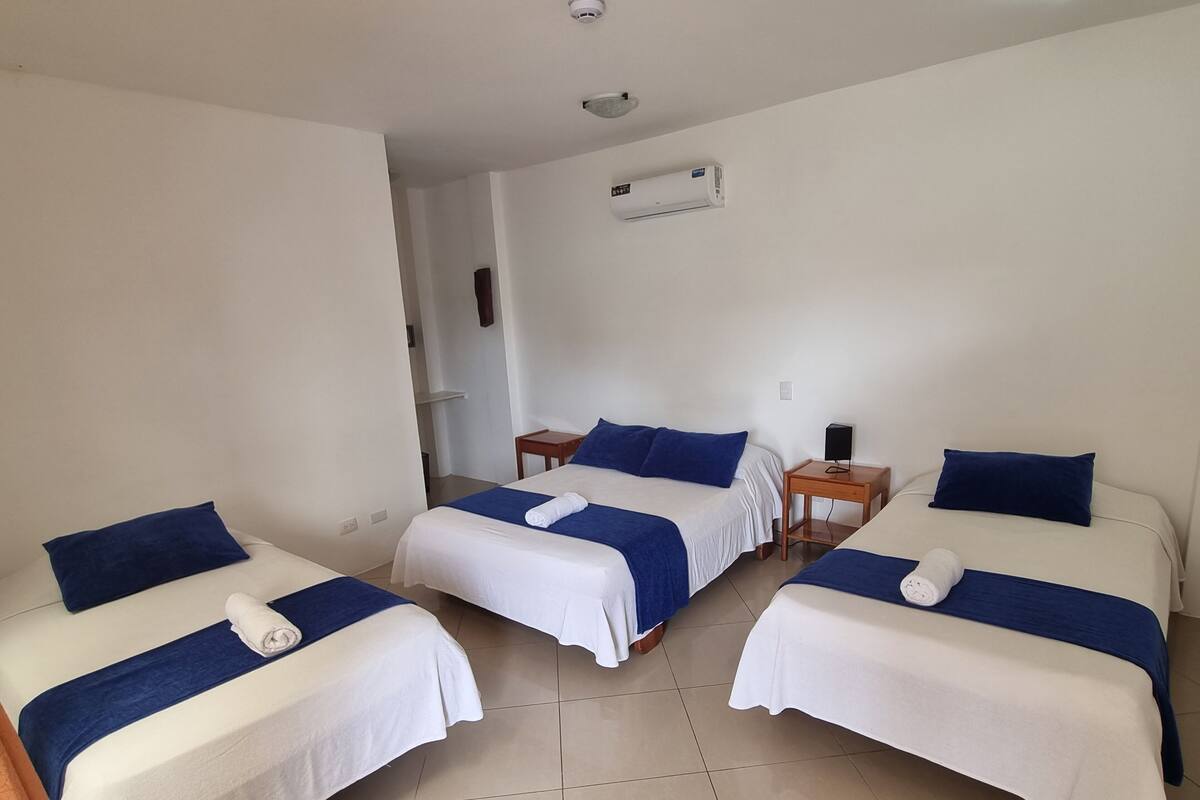 Ein Hotelzimmer mit drei Betten, jedes mit weißer Bettdecke und blauen Kissen. Am Rand steht ein kleiner Nachttisch mit Lampe und Lautsprecher.