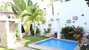 Una piscina envoltada per un jardí amb palmeres, una paret de pedra i una petita zona de terrassa.