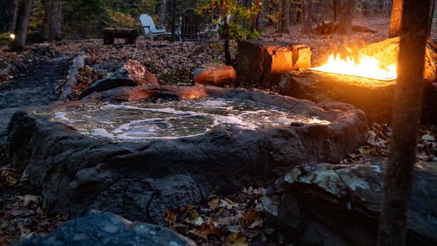 A stone fire pit surrounded by rocks and fallen leaves, with a bench and chairs in the background.