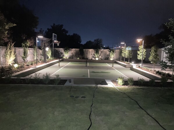 An outdoor basketball court at night with surrounding trees and a wooden fence. - A Waco cottage with private pickleball court! (Waco)