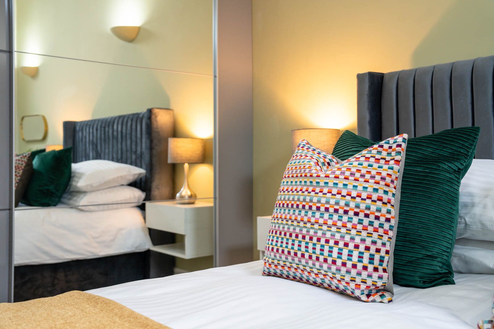 A neatly made bed with white linens, a colorful patterned pillow, a green pillow, and a dark headboard.