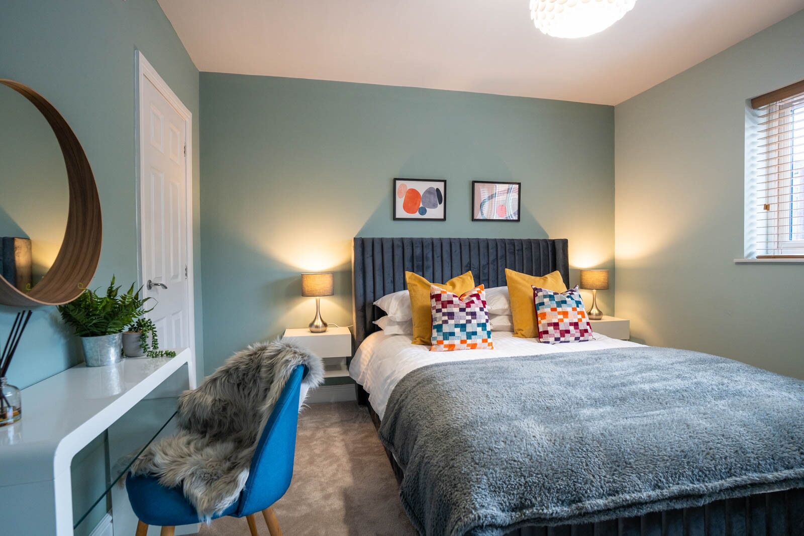 A bedroom with a bed, two bedside lamps, a mirror, a chair, and a window with blinds.