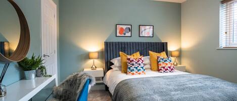 A bedroom with a bed, two bedside lamps, a mirror, a chair, and a window with blinds.