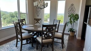 A dining area with a round table, six chairs, and a chandelier. There are large windows with a view of greenery.