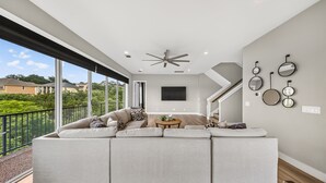Een ruime woonkamer met een grote hoekbank, een plafondventilator, een flatscreen-tv en een balkon met uitzicht op groen en omliggende huizen.