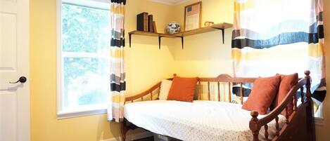 A bedroom with a wooden bed, a window with curtains, a shelf with books and a vase, and a door.