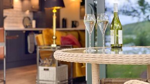 A round wicker table with two wine glasses and a bottle of wine, set against a backdrop of a modern kitchen and a view of greenery. - Waters Edge, a romantic cottage with stunning views of Talkin Tarn (Brampton)
