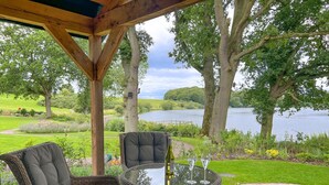 A covered outdoor seating area with wicker furniture and a glass table, overlooking a lake and lush greenery. - Waters Edge, a romantic cottage with stunning views of Talkin Tarn (Brampton)
