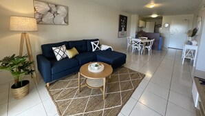 A living room with a dark blue sofa, a small round table, and a lamp. There is a kitchen in the background with white cabinets and a dining table. - City Stadium Apartment Riverfront 38 (Townsville)