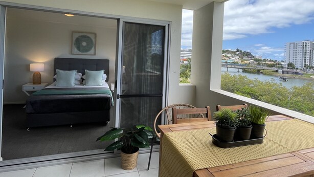 A balcony with a bed, a table with potted plants, and a view of a river and buildings. - City Stadium Apartment Riverfront 38 (Townsville)