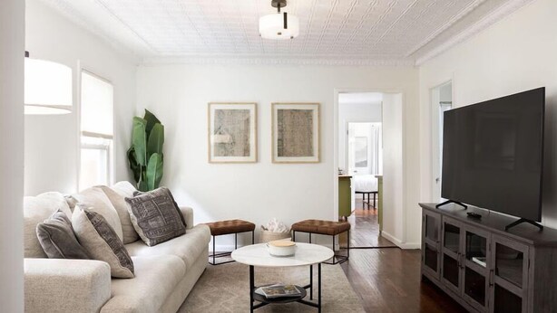 Ein Wohnzimmer mit einer Couch, einem Couchtisch und einer TV-Bank. An der Wand hängen zwei gerahmte Kunstwerke und links ist ein großes Fenster zu sehen.