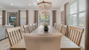 Uma sala de jantar com uma mesa de madeira comprida, cadeiras estofadas bege, um lustre e vista para o exterior através de grandes janelas.